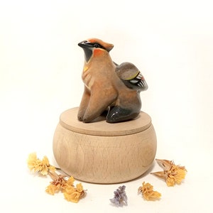 May include: A ceramic bird figurine with brown, black, and yellow accents sits on top of a wooden box with a lid. The box is surrounded by dried flowers.