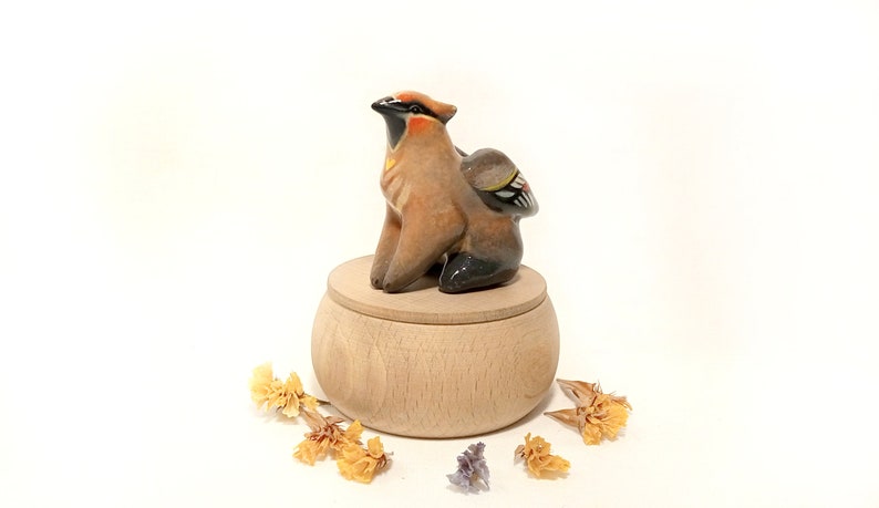 May include: A ceramic bird figurine with brown, black, and yellow accents sits on top of a wooden box with a lid. The box is surrounded by dried flowers.