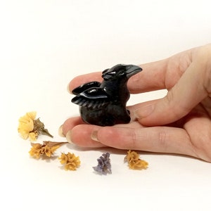 May include: A small black obsidian raven figurine with wings outstretched, held in a hand against a white background.