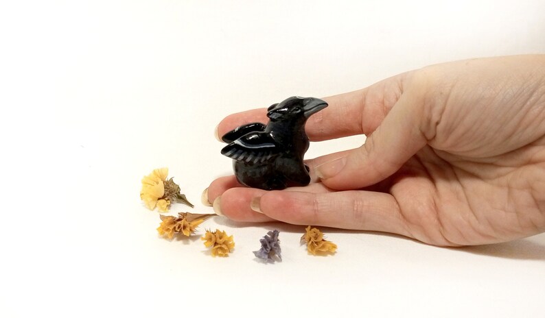 May include: A small black obsidian raven figurine with wings outstretched, held in a hand against a white background.