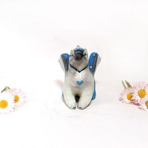 May include: A ceramic figurine of a bird with outstretched wings, painted in shades of blue, gray, and white, with a blue heart on its chest. The bird is surrounded by white daisies with yellow centers.