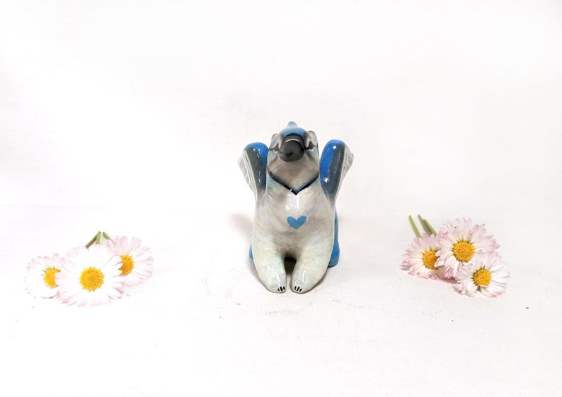 May include: A ceramic figurine of a bird with outstretched wings, painted in shades of blue, gray, and white, with a blue heart on its chest. The bird is surrounded by white daisies with yellow centers.