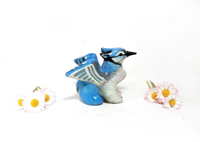 May include: A ceramic blue jay figurine with outstretched wings, painted in shades of blue, white, and gray. The bird has a black beak and is positioned on a white surface, with small white and yellow flowers on either side.