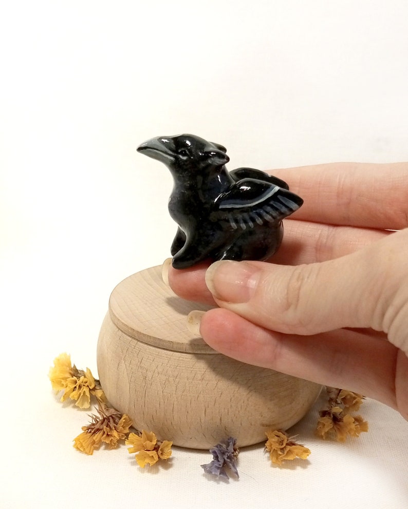 May include: A small black ceramic figurine of a mythical creature with wings. The creature has a long beak and is perched on a wooden box.