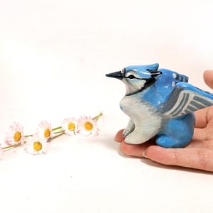 May include: A hand-painted ceramic figurine of a blue jay bird with outstretched wings. The bird is primarily blue and white, with black accents. The figurine is held in a hand, with a few daisies in the background.
