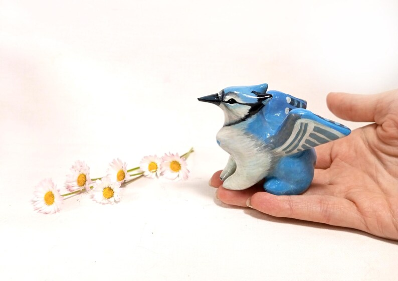 May include: A hand-painted ceramic figurine of a blue jay bird with outstretched wings. The bird is primarily blue and white, with black accents. The figurine is held in a hand, with a few daisies in the background.