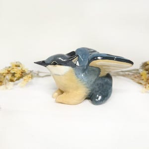 May include: A ceramic bird ornament with outstretched wings. The bird has a blue and grey body with a beige breast and a black beak. The wings are detailed with a cream and blue colour scheme. The ornament is set against a white background.
