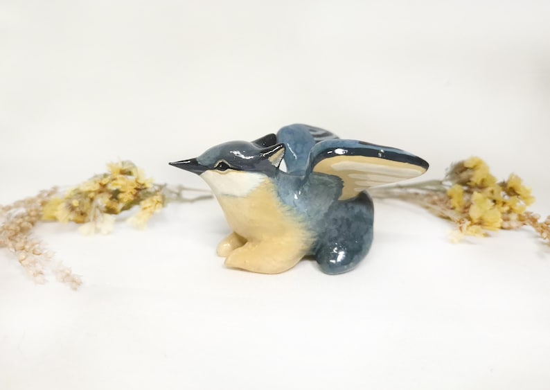 May include: A ceramic bird ornament with outstretched wings. The bird has a blue and grey body with a beige breast and a black beak. The wings are detailed with a cream and blue colour scheme. The ornament is set against a white background.