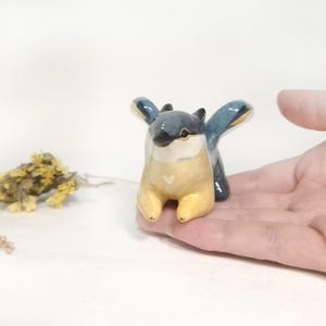 May include: A ceramic figurine of a stylized creature with blue wings, a yellow body, and a white heart detail. The figurine is held in a hand, with dried flowers in the background. The creature has a whimsical design.