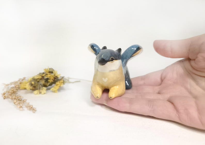 May include: A ceramic figurine of a stylized creature with blue wings, a yellow body, and a white heart detail. The figurine is held in a hand, with dried flowers in the background. The creature has a whimsical design.