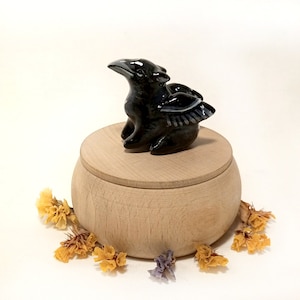 May include: A black ceramic bird figurine with wings sits on top of a wooden box. The box is round and has a lid. The bird has a long beak and is perched on the box.