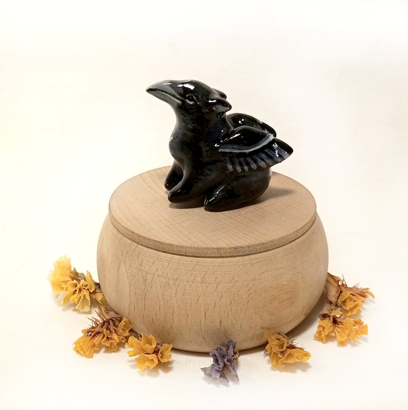 May include: A black ceramic bird figurine with wings sits on top of a wooden box. The box is round and has a lid. The bird has a long beak and is perched on the box.