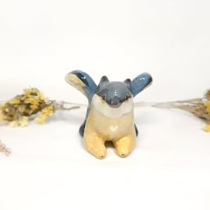 May include: A ceramic squirrel figurine with outstretched blue wings, a grey head, and a cream-coloured body. A small white heart is painted on the chest. Dried yellow flowers are on either side of the figurine.