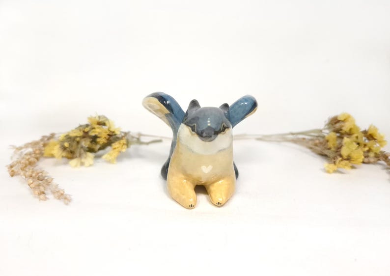 May include: A ceramic squirrel figurine with outstretched blue wings, a grey head, and a cream-coloured body. A small white heart is painted on the chest. Dried yellow flowers are on either side of the figurine.