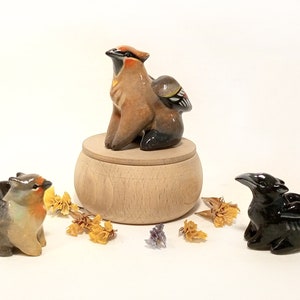 May include: Three ceramic bird figurines, each with wings. The first is brown and orange with a black head and is perched on a wooden box. The second is brown and gray with a red breast and the third is black with a long beak.