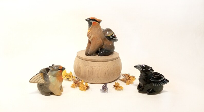 May include: Three ceramic bird figurines, each with wings. The first is brown and orange with a black head and is perched on a wooden box. The second is brown and gray with a red breast and the third is black with a long beak.