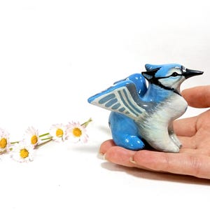 May include: A ceramic figurine of a blue jay with white and black accents. The bird is perched on a hand, with a sprig of daisies in the background. The figurine is approximately 3 inches in length.