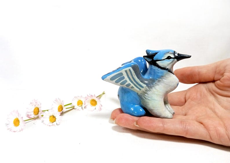 May include: A ceramic figurine of a blue jay with white and black accents. The bird is perched on a hand, with a sprig of daisies in the background. The figurine is approximately 3 inches in length.