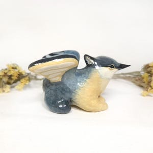 May include: A ceramic bird ornament with outstretched wings. The bird has a blue-grey body, tan chest, and a black beak. The wings are blue and tan. Dried yellow flowers are in the background.