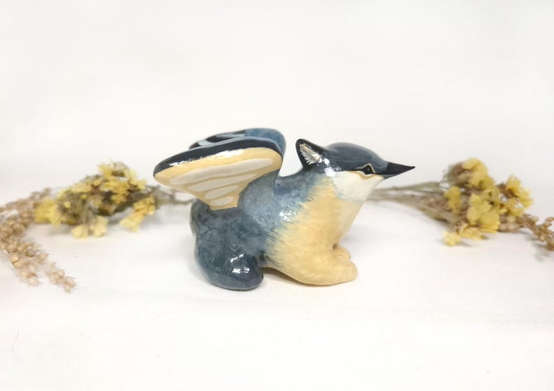May include: A ceramic bird ornament with outstretched wings. The bird has a blue-grey body, tan chest, and a black beak. The wings are blue and tan. Dried yellow flowers are in the background.