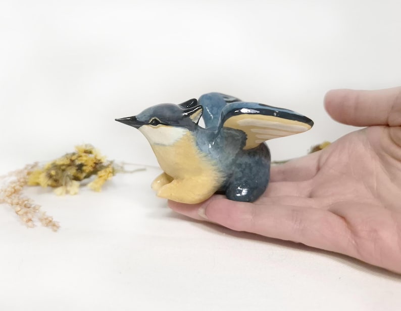 May include: A ceramic bird ornament with open wings. The bird features a blue and grey body, yellow breast, and black beak. The figurine is held in a hand against a white backdrop. Dried flowers are in the background.