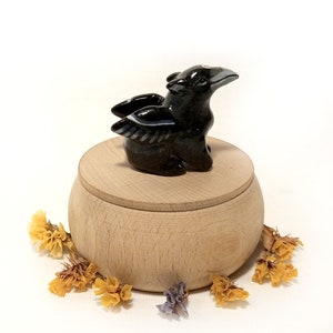 May include: A black ceramic crow figurine with white markings sits on top of a wooden box with a lid. The box is round and has a natural wood finish.