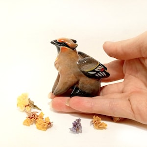 May include: A small ceramic figurine of a brown and black bird with a red and white wing. The bird is sitting on a hand.