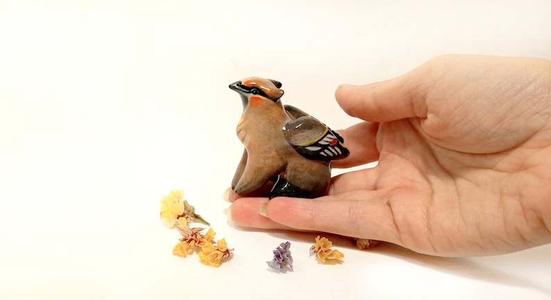 May include: A small ceramic figurine of a brown and black bird with a red and white wing. The bird is sitting on a hand.