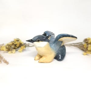 May include: A ceramic bird figurine in blue and tan. The bird has outstretched wings and a detailed beak. Dried yellow flowers are on either side of the bird. The figurine is a decorative item.