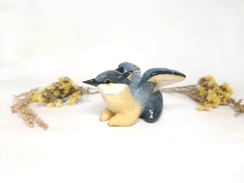 May include: A ceramic bird figurine in blue and tan. The bird has outstretched wings and a detailed beak. Dried yellow flowers are on either side of the bird. The figurine is a decorative item.