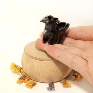 May include: A small black ceramic dragon figurine with a white heart on its chest. The dragon is perched on a wooden box with a lid.