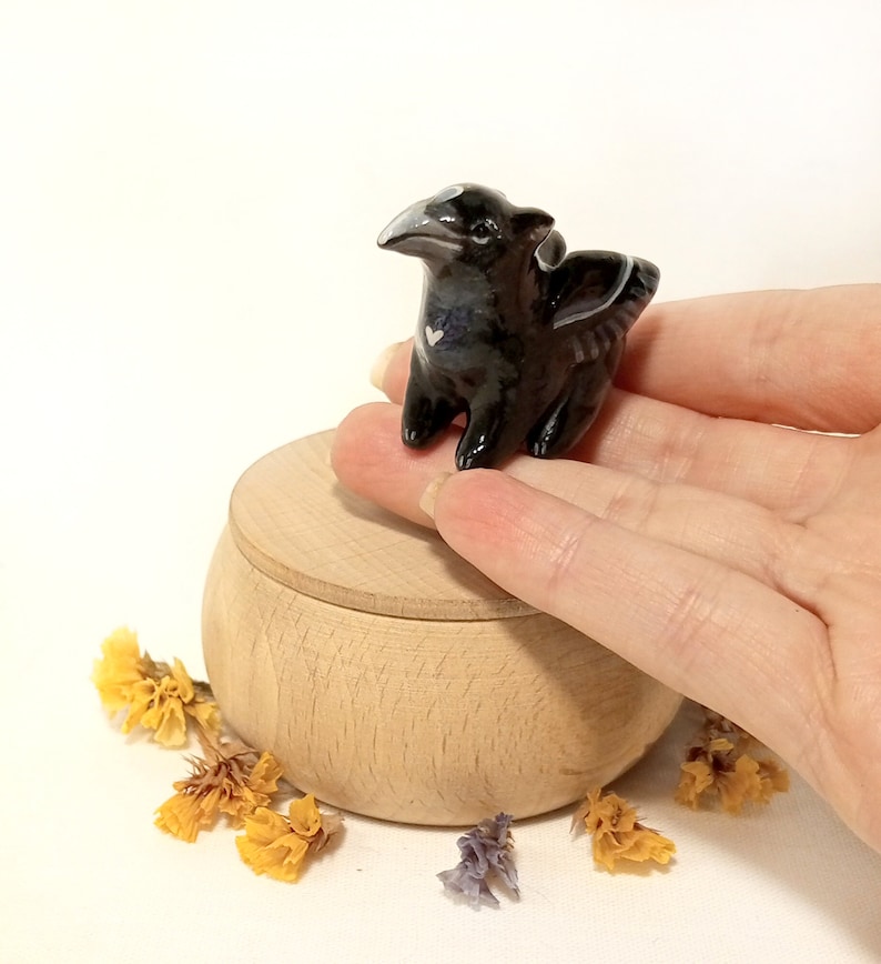 May include: A small black ceramic dragon figurine with a white heart on its chest. The dragon is perched on a wooden box with a lid.