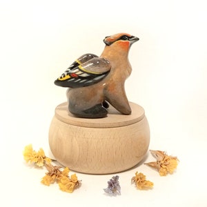 May include: A ceramic bird figurine with brown, black, yellow, and red painted details, perched on a wooden box with a lid. The bird has its wings spread out and is looking to the right. The box is made of light wood and has a smooth surface.
