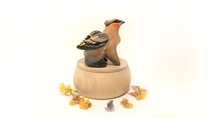 May include: A ceramic bird figurine with brown, black, yellow, and red painted details, perched on a wooden box with a lid. The bird has its wings spread out and is looking to the right. The box is made of light wood and has a smooth surface.
