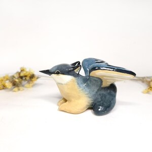 May include: A ceramic bird figurine with outstretched wings. The bird has a blue and grey body with a cream-coloured chest and wings. The beak is black. The figurine is a decorative item.