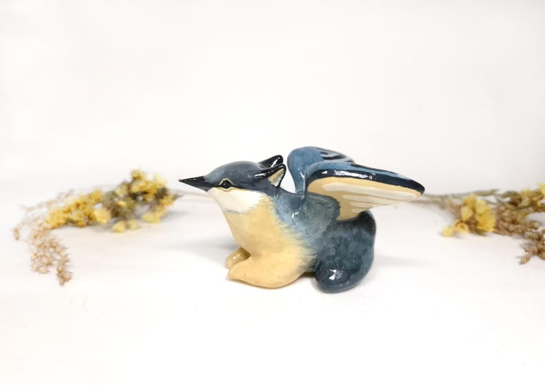 May include: A ceramic bird figurine with outstretched wings. The bird has a blue and grey body with a cream-coloured chest and wings. The beak is black. The figurine is a decorative item.