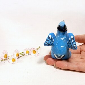 May include: A hand-painted blue bird figurine with black accents and white details on the wings and tail. The bird is perched on a hand, with a sprig of daisies in the background. The figurine is a decorative item.