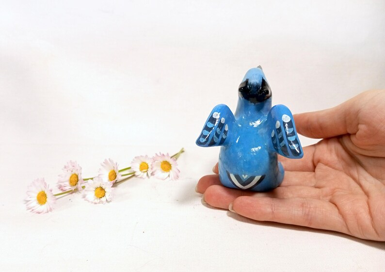 May include: A hand-painted blue bird figurine with black accents and white details on the wings and tail. The bird is perched on a hand, with a sprig of daisies in the background. The figurine is a decorative item.