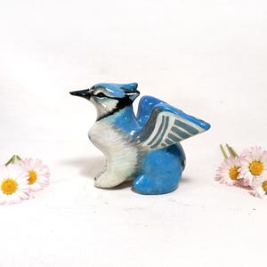 May include: A ceramic figurine of a blue jay with outstretched wings. The bird is primarily blue and white, with black accents on its beak and head. Two small bouquets of daisies with white petals and yellow centers are on either side of the bird.