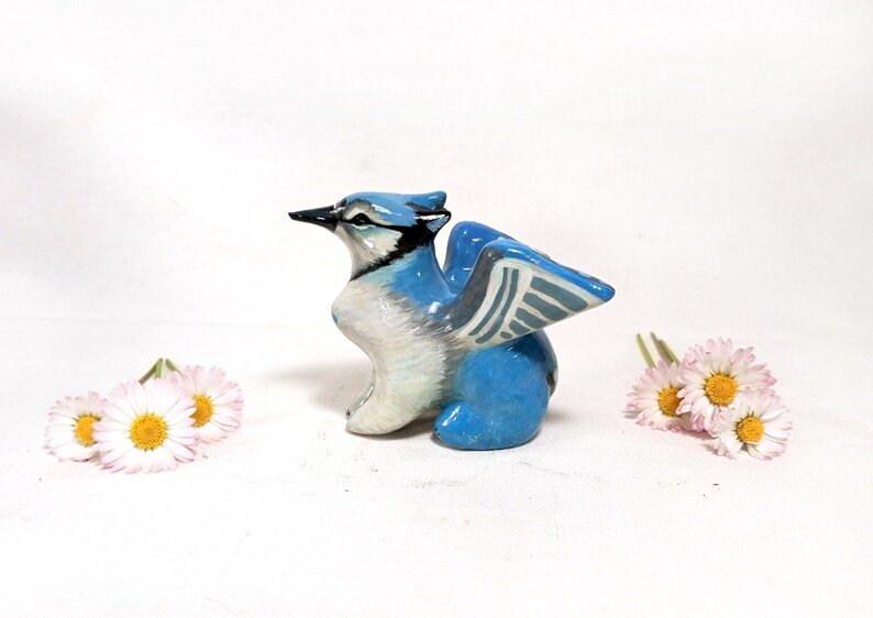 May include: A ceramic figurine of a blue jay with outstretched wings. The bird is primarily blue and white, with black accents on its beak and head. Two small bouquets of daisies with white petals and yellow centers are on either side of the bird.