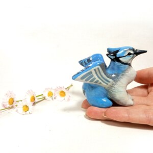 May include: A hand-painted, ceramic blue jay figurine with a detailed design. The bird has blue, white, and black coloring, with its wings slightly spread. A few white and yellow daisies are in the background.