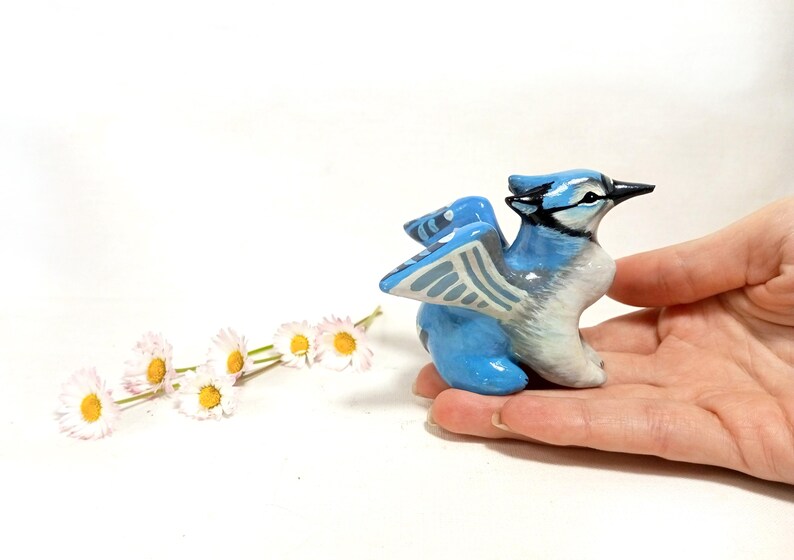 May include: A hand-painted, ceramic blue jay figurine with a detailed design. The bird has blue, white, and black coloring, with its wings slightly spread. A few white and yellow daisies are in the background.