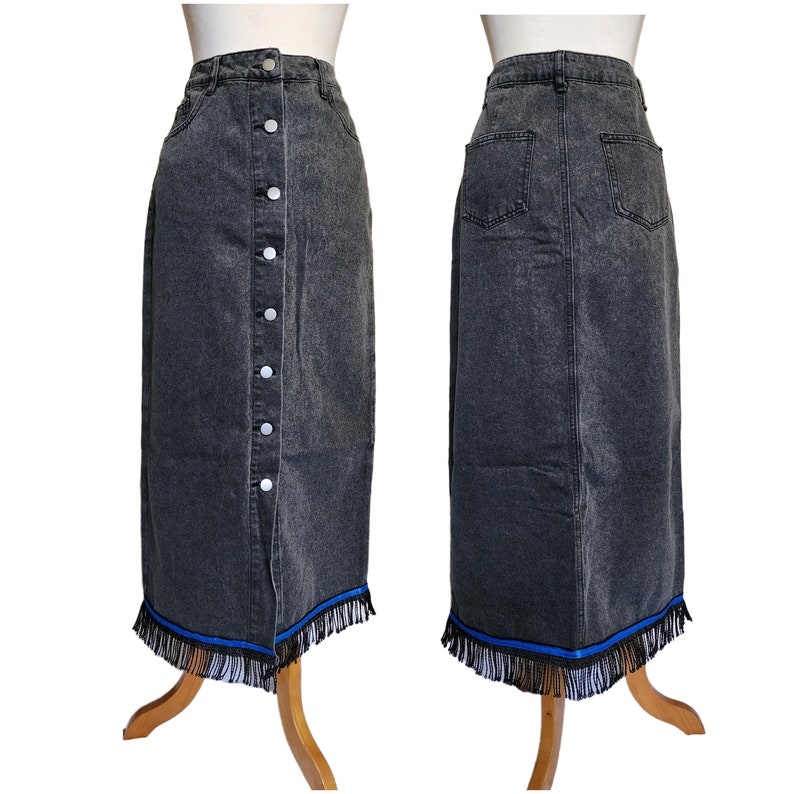May include: A dark gray denim skirt with a button-up front and a fringed hemline. The skirt has a high waist and a straight silhouette. The hemline features a blue ribbon detail and black fringe.