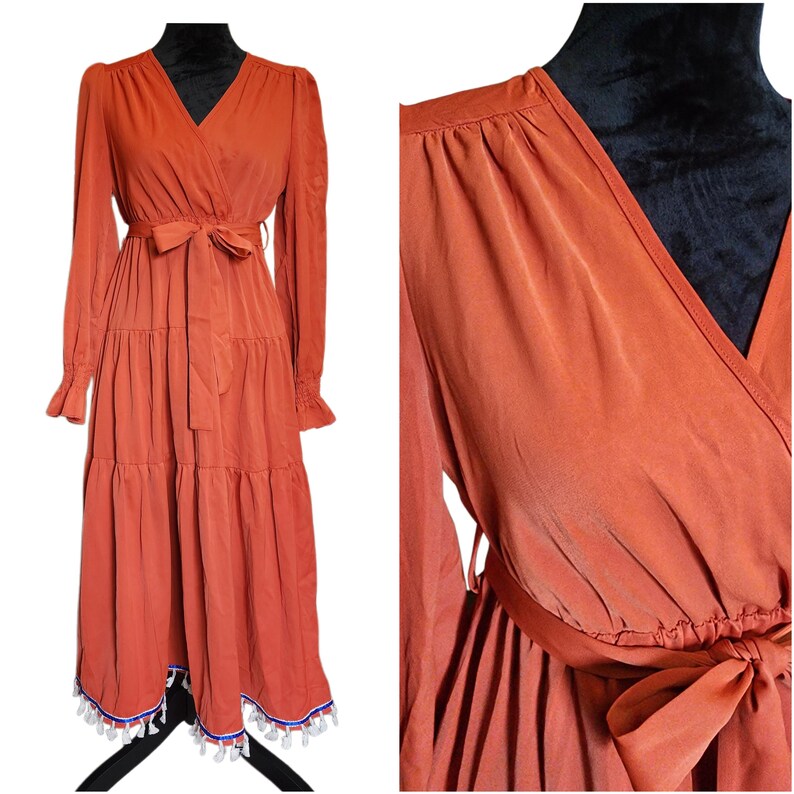 May include: Rust-colored wrap dress with long sleeves and a tiered skirt. The dress features a V-neck, a waist tie, and ruffled cuffs. The hemline is adorned with white tassels and blue and white trim. The dress is made of a flowing fabric.