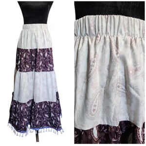 May include: A long, tiered skirt with a paisley pattern in shades of cream and dark purple. The skirt has an elastic waistband and is trimmed with tassels along the bottom hem. The skirt is displayed on a mannequin.