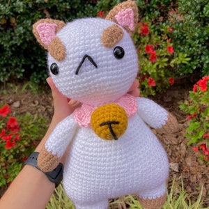May include: White crocheted cat plush toy with brown and pink accents. The cat has a pink collar with a gold bell.