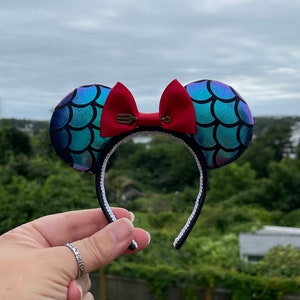 Może przedstawiać: Para uszu Minnie Mouse w stylu syreny, w kolorze niebieskim i zielonym, z czerwoną kokardką i zawieszką w kształcie widelca. Uszy są wykonane z błyszczącego materiału i mają białe opaski.
