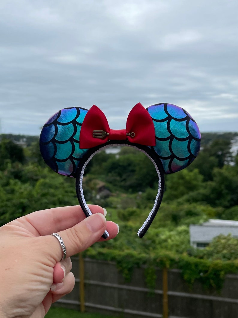 Może przedstawiać: Para uszu Minnie Mouse w stylu syreny, w kolorze niebieskim i zielonym, z czerwoną kokardką i zawieszką w kształcie widelca. Uszy są wykonane z błyszczącego materiału i mają białe opaski.