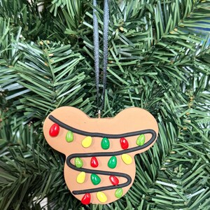 May include: A brown, Mickey Mouse-shaped Christmas ornament with colorful lights. The ornament has a black string for hanging and is set against a backdrop of green Christmas tree branches.