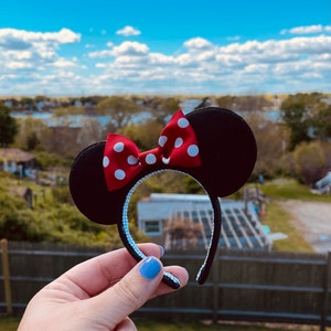 May include: Black Minnie Mouse ears headband with a red and white polka dot bow. The headband is held in a hand against a blurred background of a blue sky and green trees.